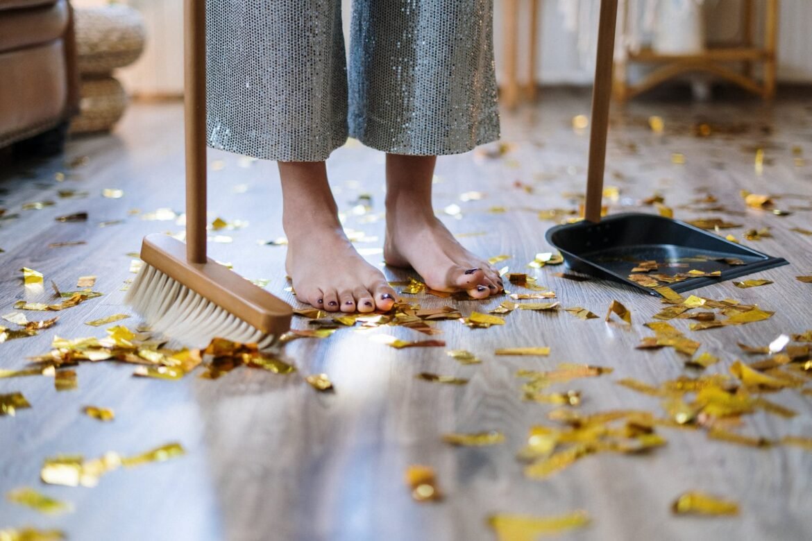 Quoi de neuf : du nettoyage ! Une femme (dont on ne voit que les pieds), passe le balai chez elle pour nettoyer un sol plein de confettis